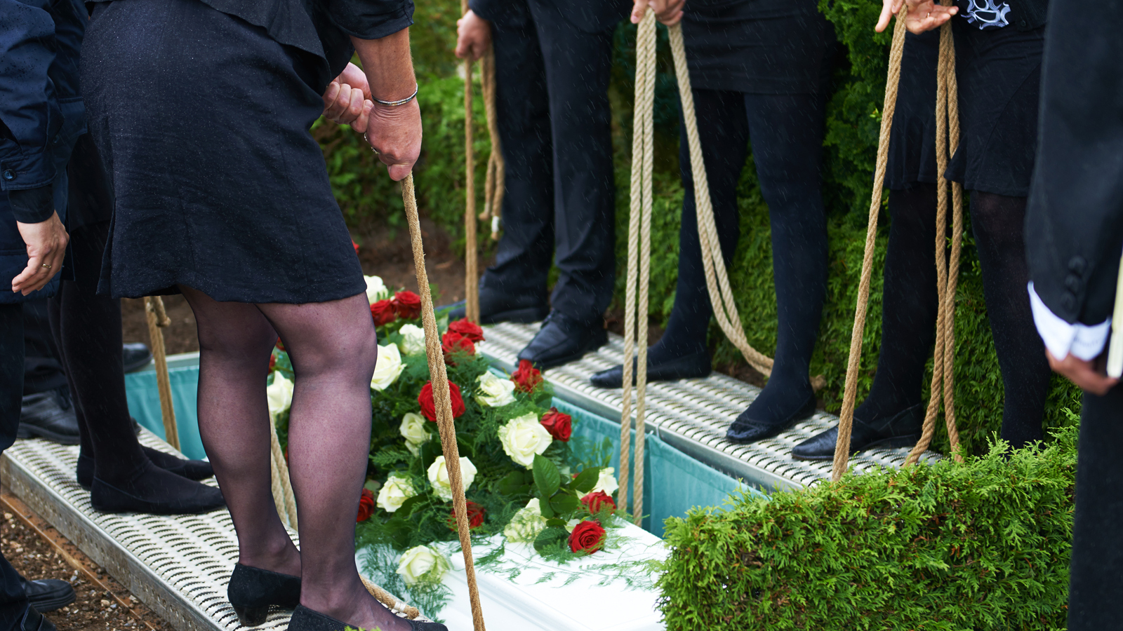Teilnehmer einer Trauerfeier halten Seile, um Blumenschmuck auf einen Sarg in einer Grabstelle abzusenken.